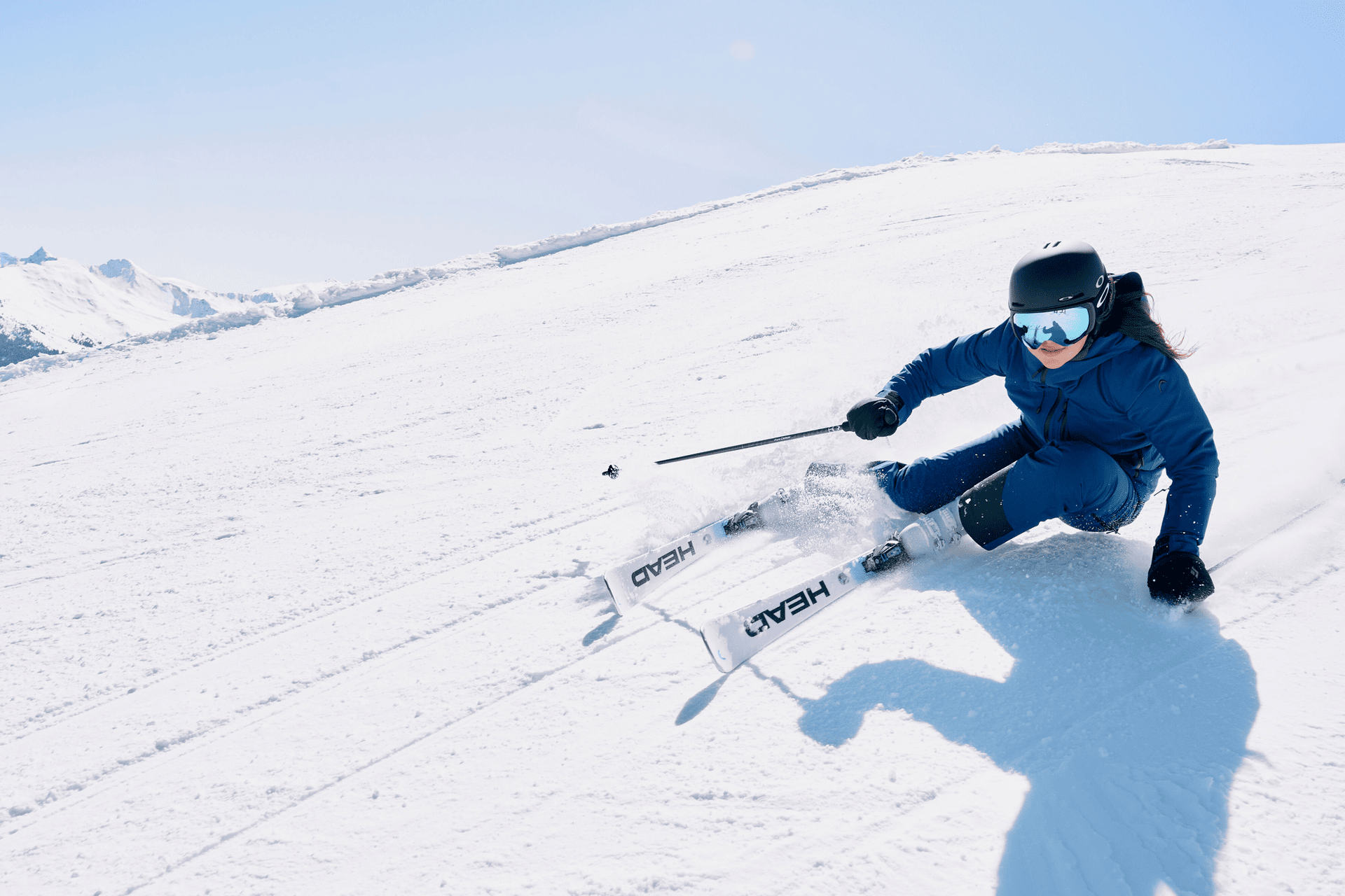 Skier in blue outfit and goggles expertly turns on a sunny snow-covered slope, casting a shadow. Snow-capped mountains are visible in the background.