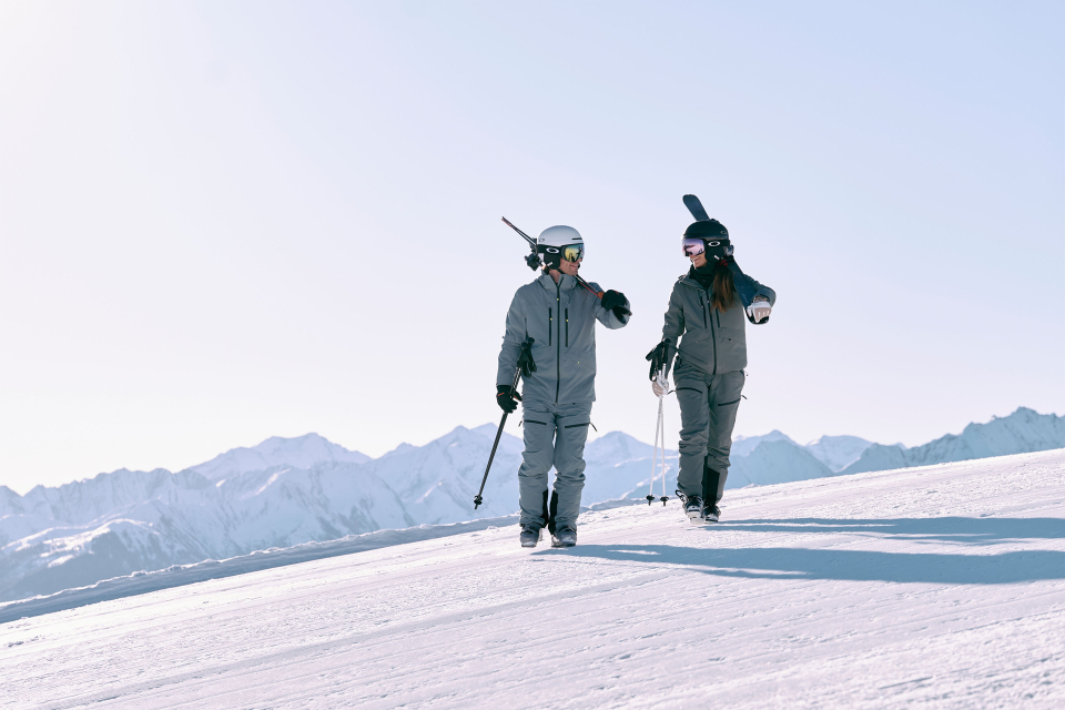 Two skiers in gray outfits walk on a snowy slope, bathed in sunlight. One carries skis, the other a snowboard. Snowy mountains form the background.