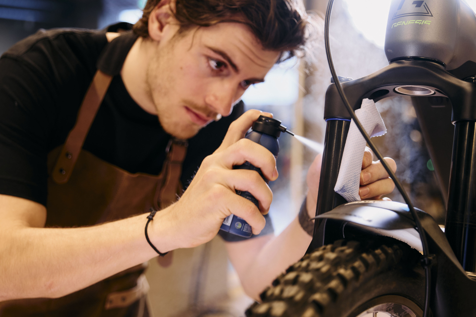 A focused man in an apron cleans a motorcycle fork with a spray can and cloth, conveying a meticulous and attentive atmosphere in a workshop setting.