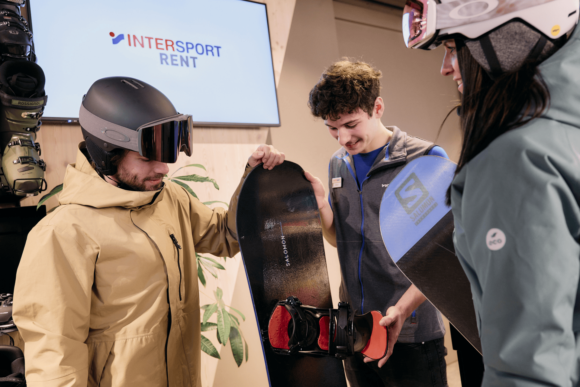 Two people in ski gear examine snowboards with a store employee in front of an "INTERSPORT RENT" sign. The atmosphere is friendly and engaging.