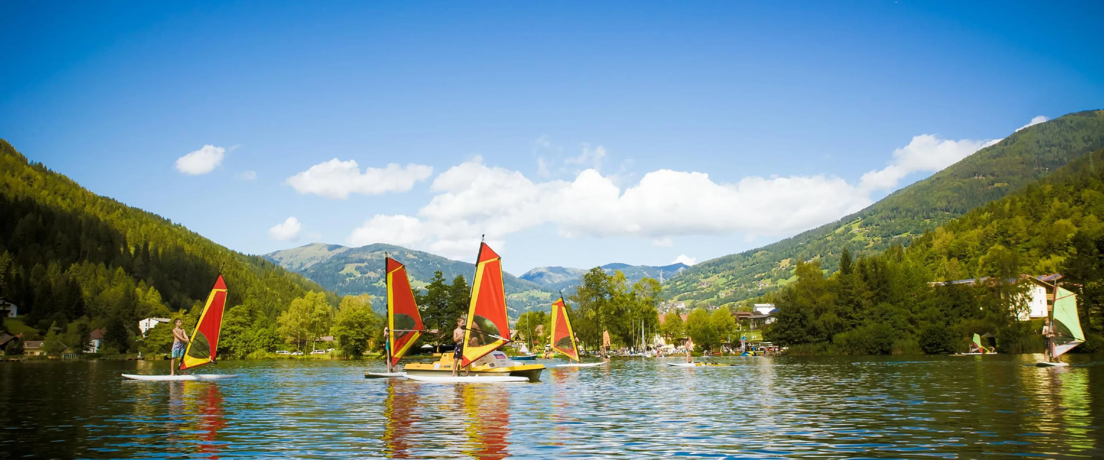 Sailboats with red and orange sails glide on a serene lake, surrounded by lush green hills under a bright blue sky and fluffy white clouds.