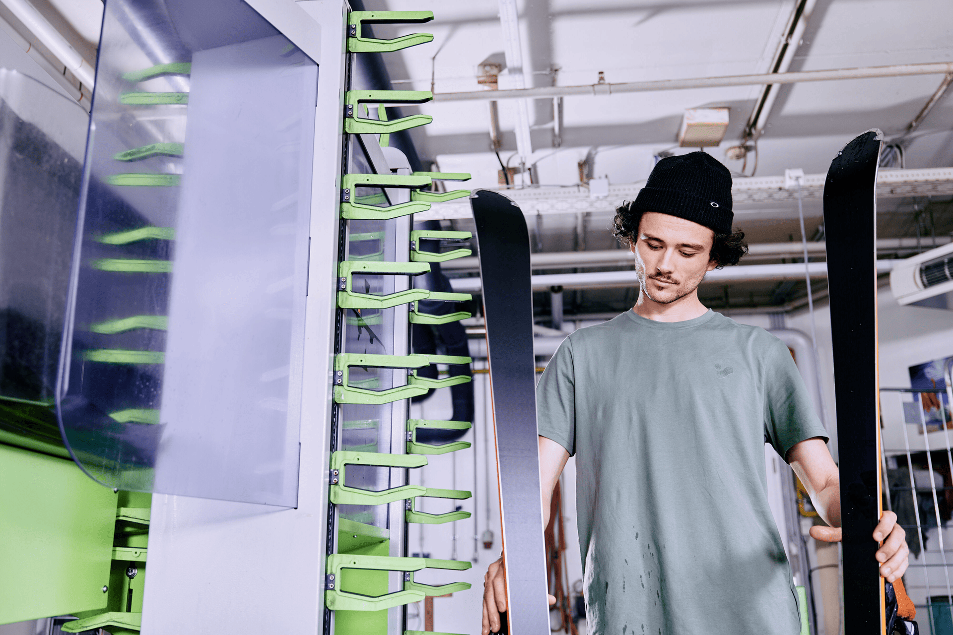 A person in a beanie and T-shirt inspects skis in a workshop. They hold the skis upright, surrounded by green equipment, with a focused expression.