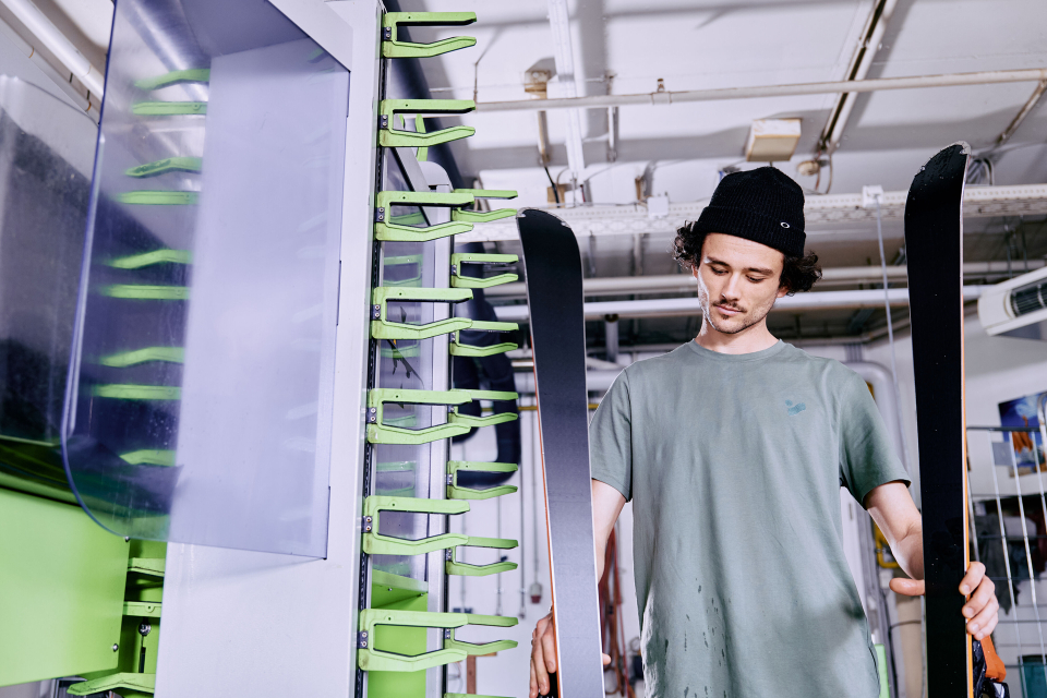 Eine Person mit Mütze und T-Shirt inspiziert Skier in einer Werkstatt. Sie hält die Skier aufrecht, umgeben von grüner Ausrüstung, mit konzentriertem Gesichtsausdruck.