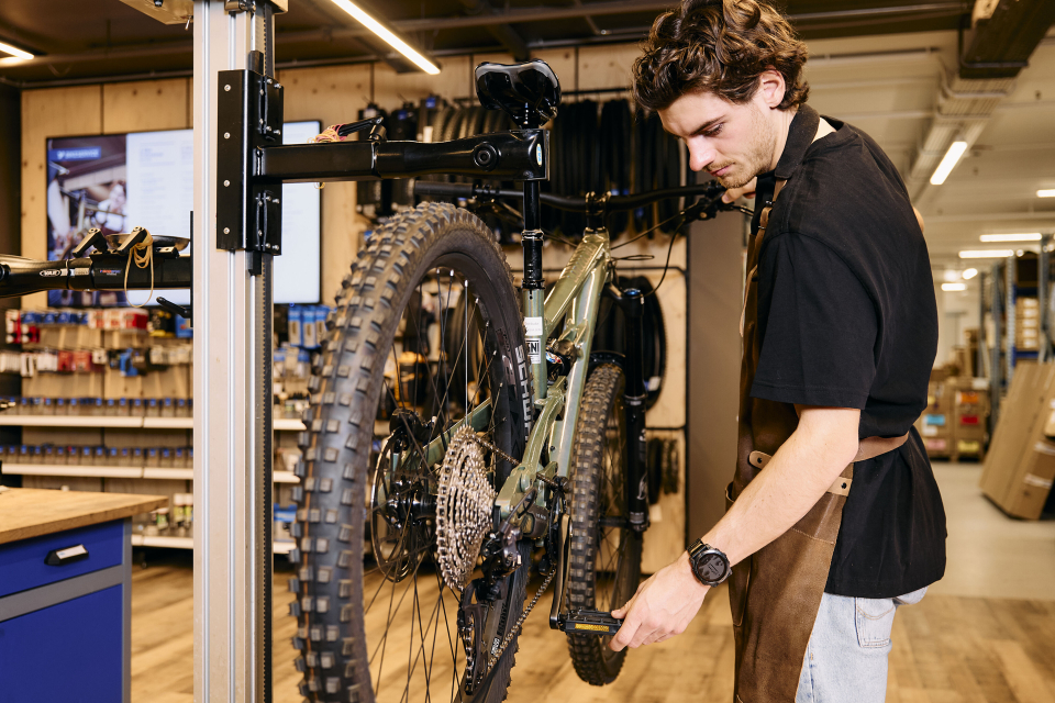 A bike mechanic in a workshop focuses intently on adjusting a mountain bike's pedal. Shelves with parts and tools are visible in the background.