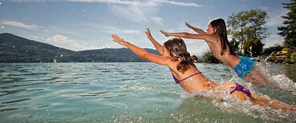 Zwei Kinder springen an einem sonnigen Tag fröhlich in einen ruhigen See, im Hintergrund sind Berge und Bäume zu sehen. Klares Wasser spritzt um sie herum.