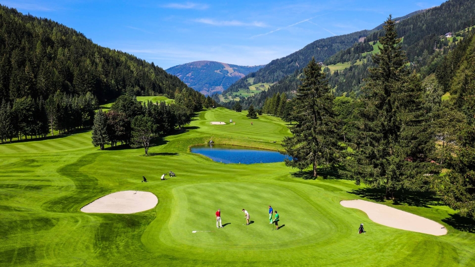 Golfers play on a lush, green course surrounded by tall trees and mountains. A small pond reflects the blue sky. The scene is peaceful and expansive.