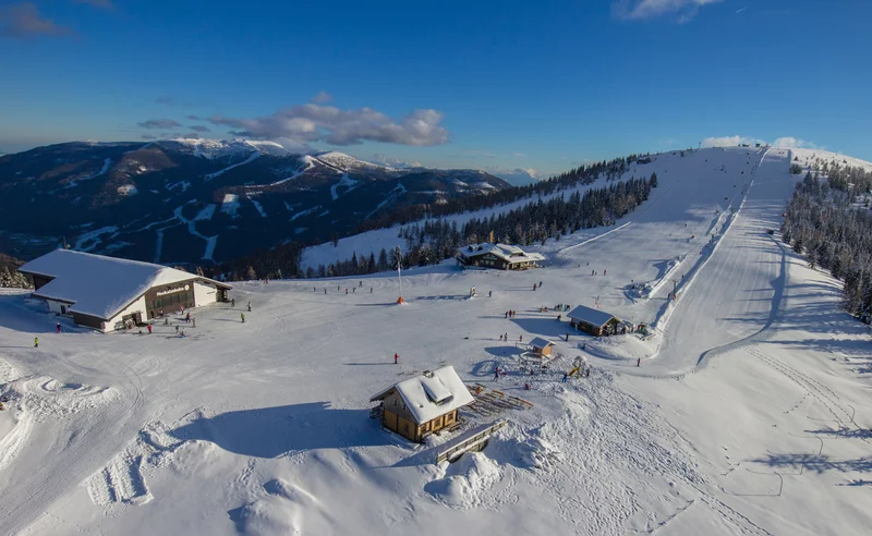 Luftaufnahme eines verschneiten Skigebiets mit einer breiten Piste, Skifahrern und gemütlichen Hütten. Umgeben von Kiefern und fernen Bergen unter einem strahlend blauen Himmel.