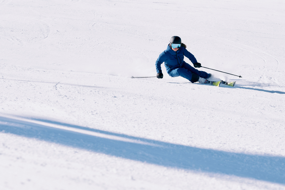 A skier dressed in blue, wearing goggles and a helmet, elegantly carves through fresh snow on a bright, sunny day, creating a dynamic motion effect.