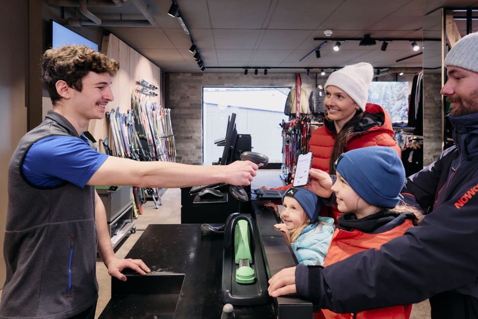 A cheerful sales associate hands a receipt to a smiling family, including a man, woman, and two kids, in a cozy ski shop filled with gear.