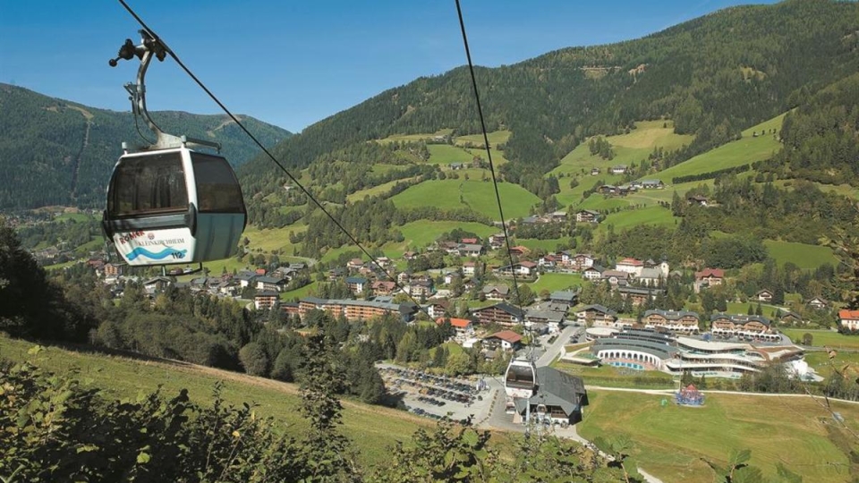 A cable car ascends over a picturesque village nestled in lush green hills. The scene is sunny and peaceful, with mountains in the background.