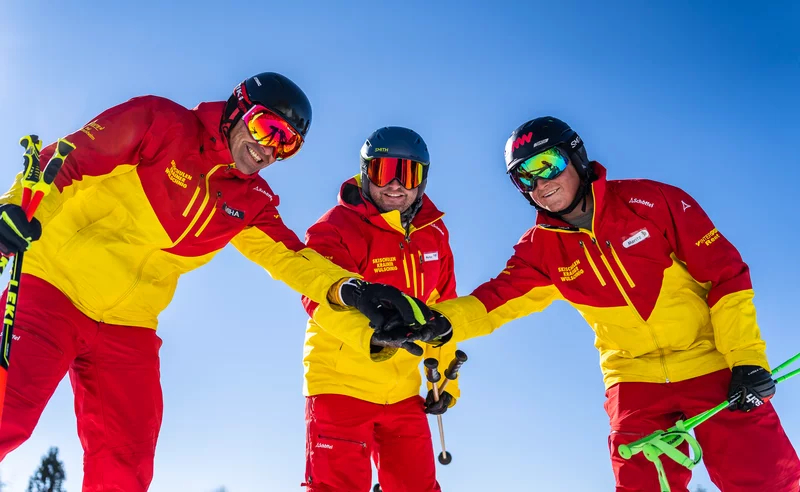 Three skiers in red and yellow suits, wearing helmets and goggles, smiling and fist-bumping on a sunny ski slope, conveying teamwork and joy.